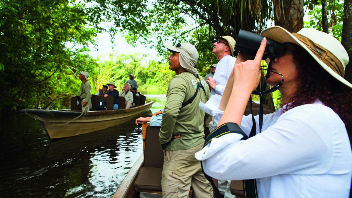 Tourists on a boat observing wildlife in a lush, green tropical environment. One person uses binoculars to watch, while others look on, surrounded by dense foliage.
