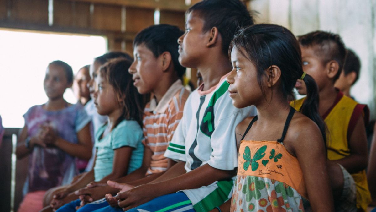 Children sitting closely together in a wooden classroom, engaged and attentive. A diverse group, featuring varying hairstyles and clothing, with some wearing bright colors. Sunlight filters through the window, illuminating their faces.