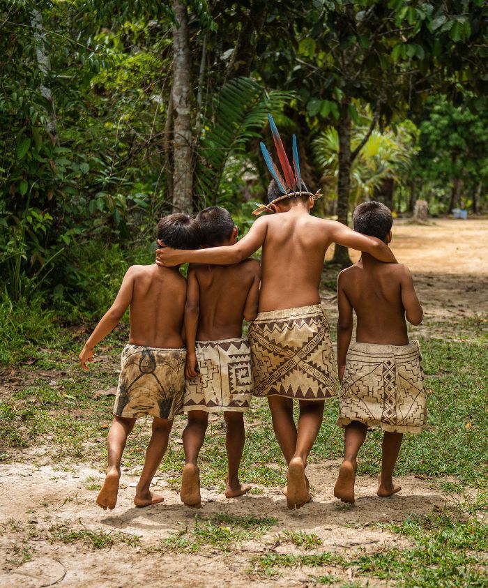 Four young boys walk together down a dirt path in a lush forest. They are wearing traditional skirts with patterns and one boy has colourful feathers in his hair. The boys appear happy, with their arms around each other.