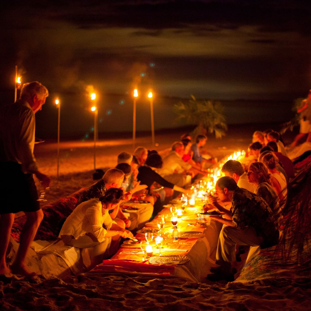 A festive beach dinner party at night, with guests seated at a long table illuminated by candles and tiki torches. The scene features people enjoying their meals, surrounded by a serene ocean backdrop under a starry sky.