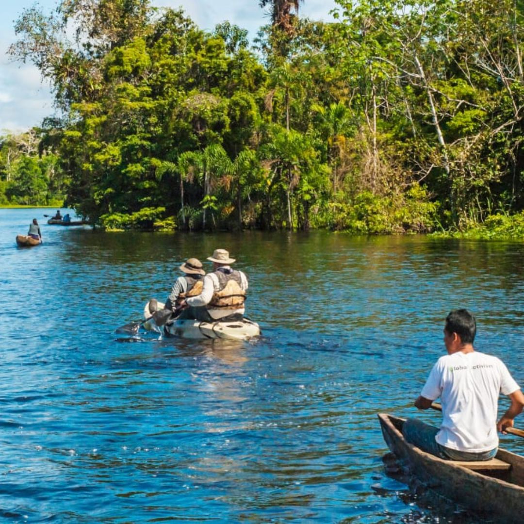 Two individuals in canoes paddle along a calm river surrounded by lush greenery. In the background, additional canoeists navigate through the vibrant landscape, with trees reflecting in the water. A lone figure sits in a wooden canoe nearby, observing.