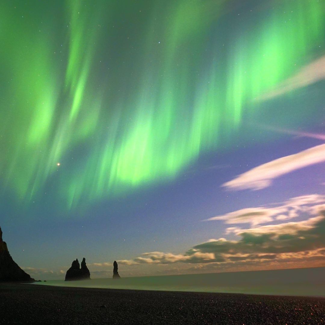 Vibrant green auroras dance across the night sky above a rocky beach, with silhouettes of jagged rock formations in the foreground and soft clouds illuminated by the lights.