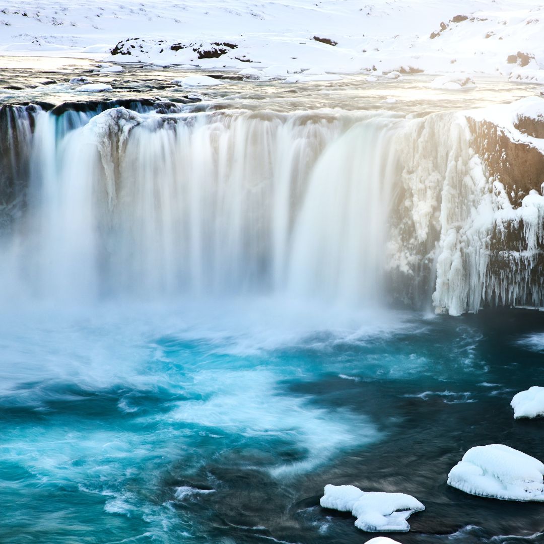 A majestic waterfall cascades over rocky cliffs into a vibrant turquoise river, surrounded by snow-covered terrain. Sunlight glimmers off the water, creating a serene winter landscape.