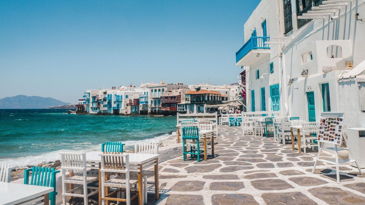 A sunlit coastal pathway lined with white buildings featuring blue accents. Seaside tables and chairs are set up along the water's edge, with gentle waves lapping at the shore. In the background, charming colorful houses reflect traditional architecture.