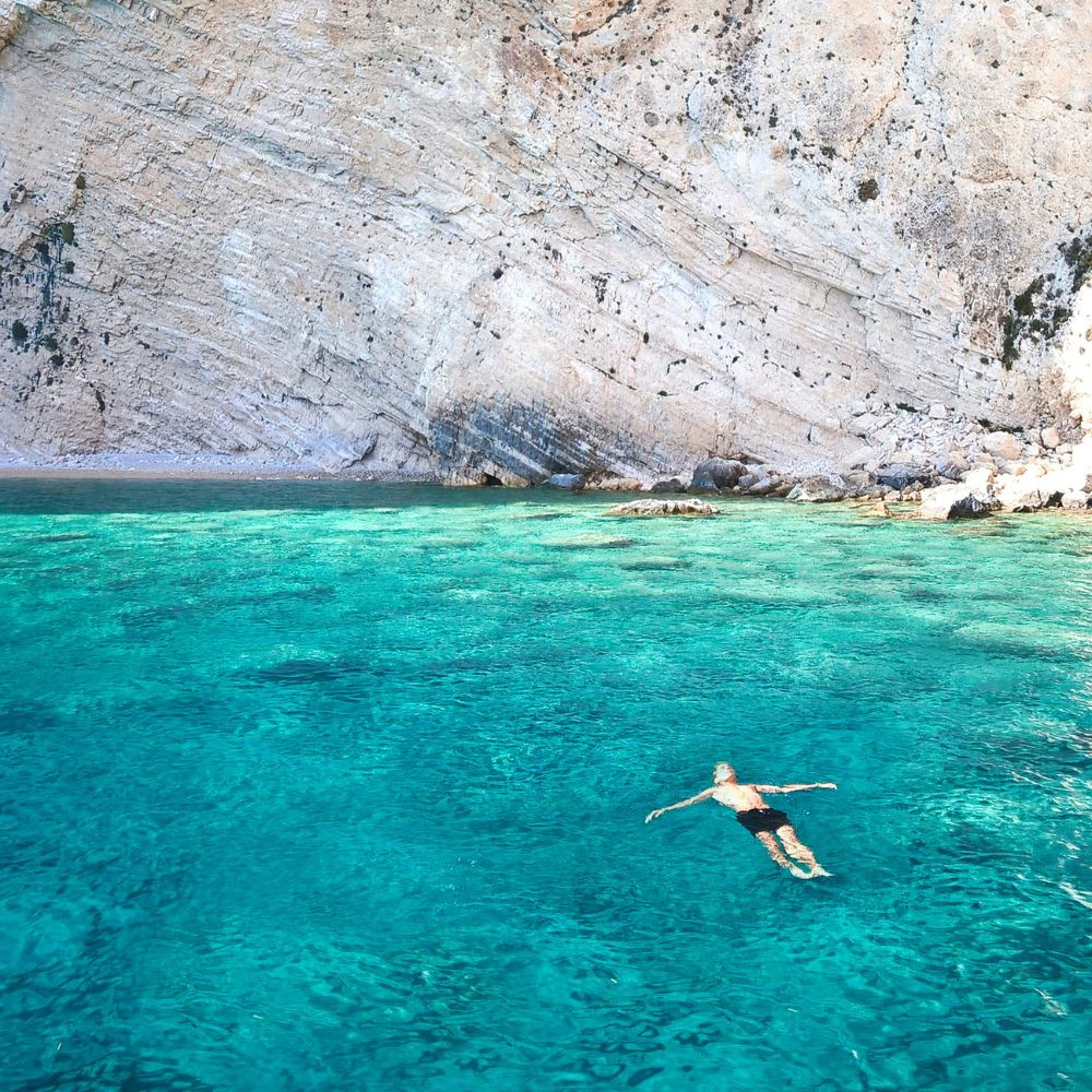 A person floats serenely on clear turquoise water near a rocky shore. The sunlight reflects off the surface, highlighting the vibrant blue tones of the sea.