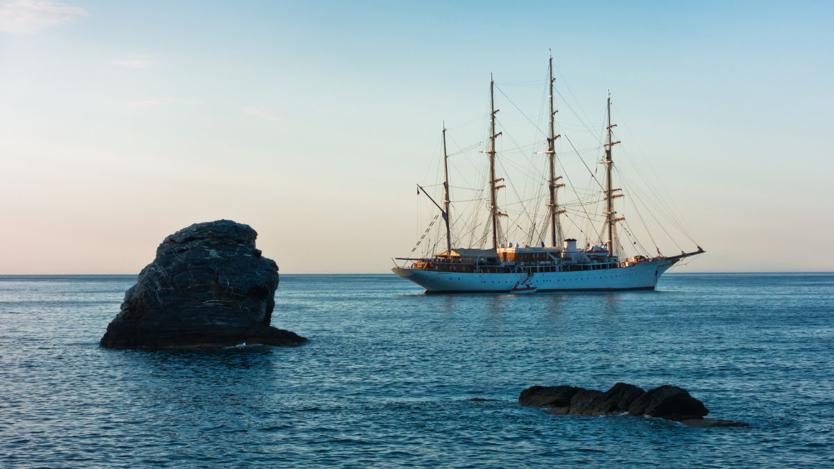 A tall ship with multiple masts floats on calm waters near a rocky outcrop, under a soft blue sky.