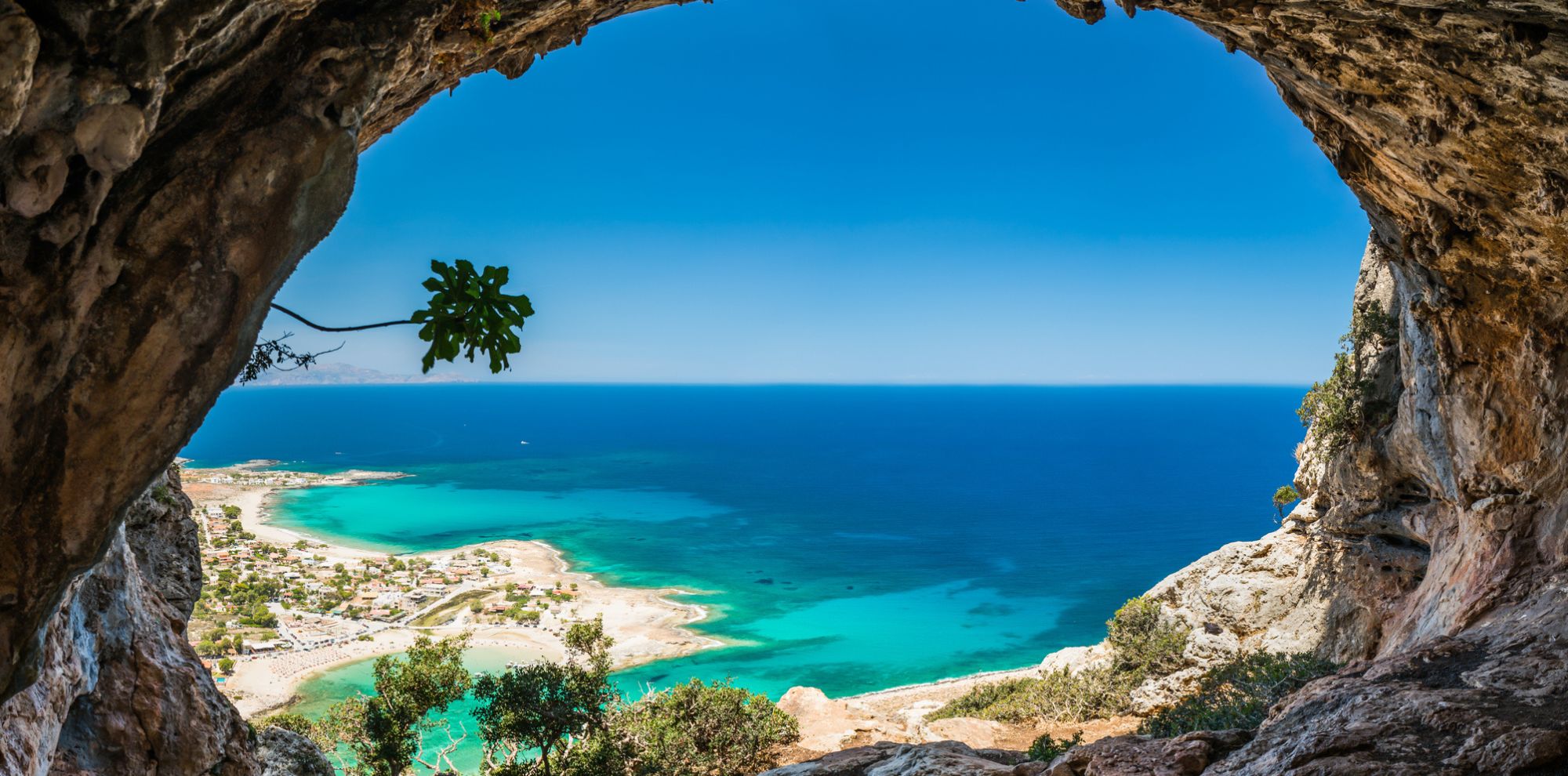 A breathtaking view from inside a cave, showcasing a vibrant blue sea and a coastal village with sandy shores under a clear sky. The lush greenery frames the scene.