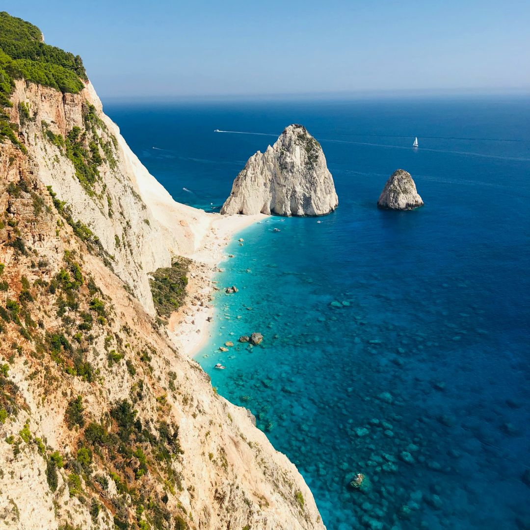 A scenic coastal view featuring steep cliffs, clear turquoise waters, and two rocky islets in the distance. A small sailboat is visible on the horizon.