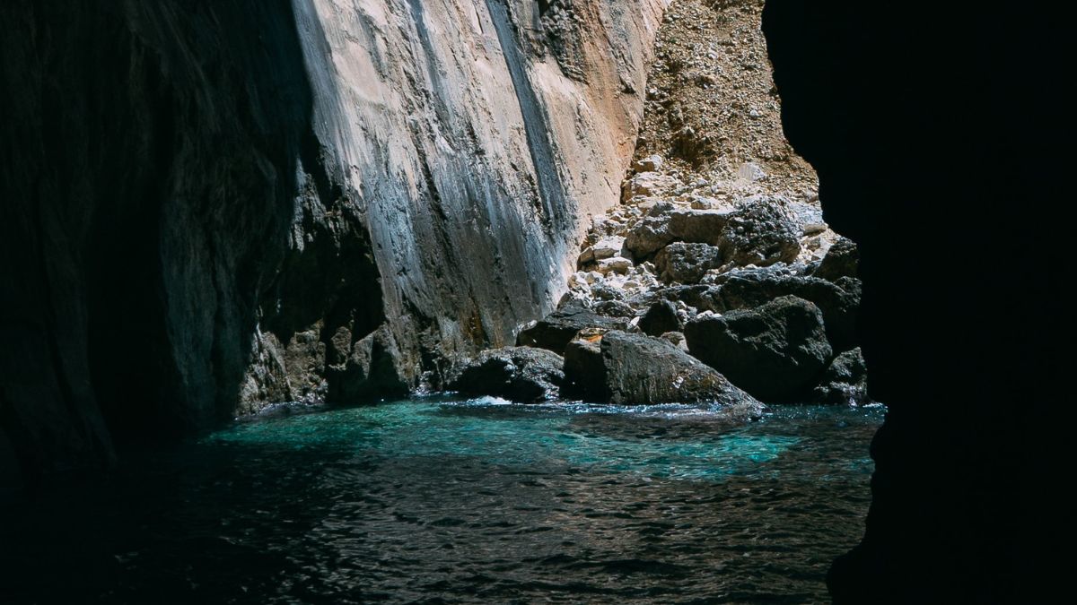 Sunlight illuminates a rocky cave entrance, revealing turquoise waters and textured stone walls. The scene combines shadows and light, highlighting the natural beauty of the coastal landscape.