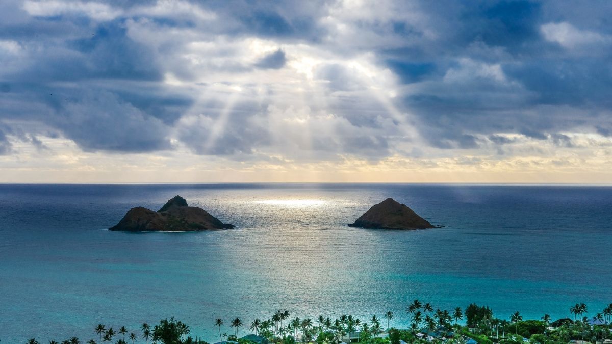 Dual islands emerge from shimmering waters under a dramatic sky, with rays of sunlight breaking through clouds above. The scene captures the tranquil beauty of the ocean and shoreline.