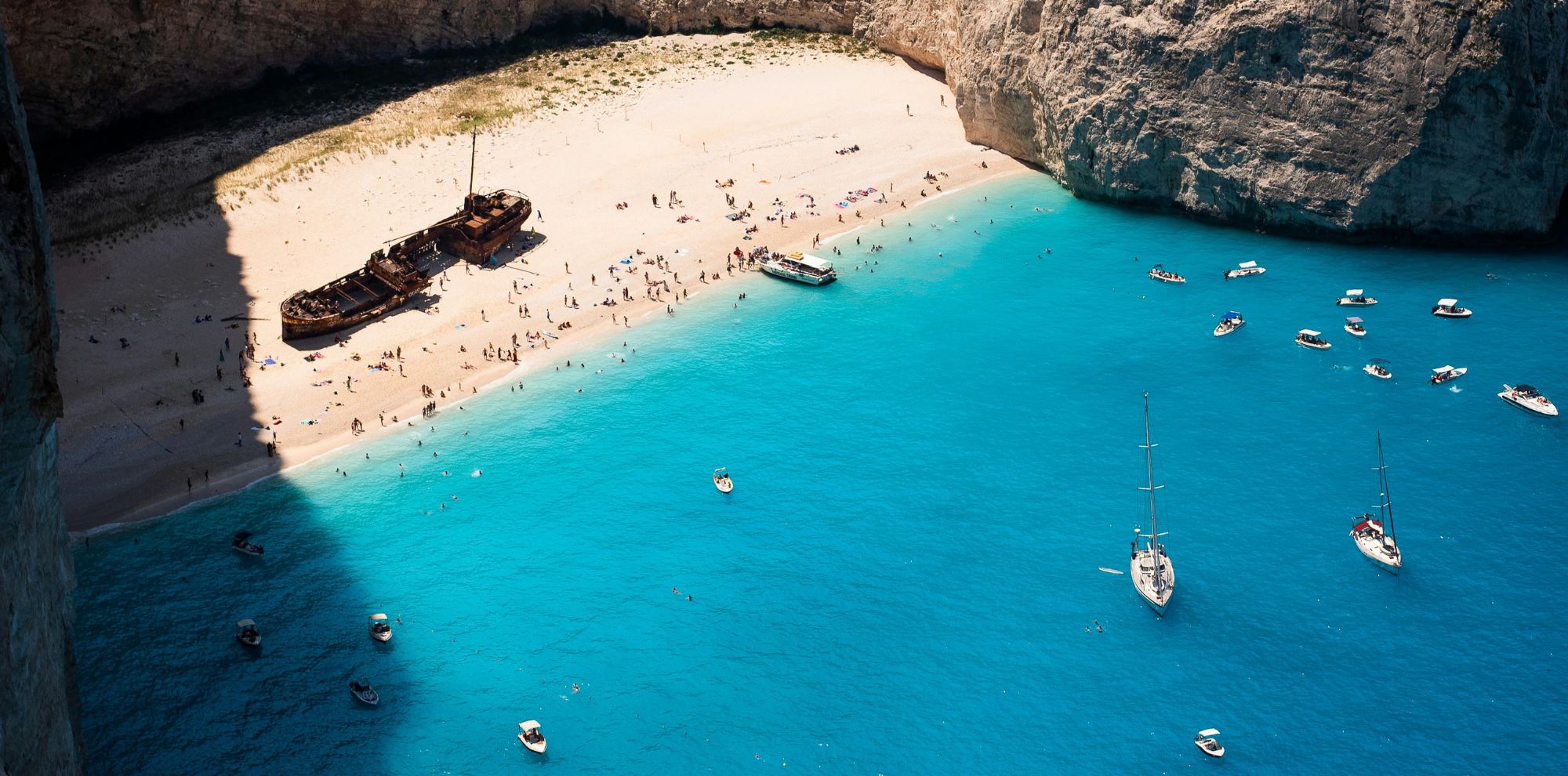 A stunning beach with crystal-clear turquoise water and a shipwreck resting on the shore. Sunbathers are scattered across the sandy beach, while boats are anchored nearby. Tall cliffs surround the area, creating a picturesque scene.