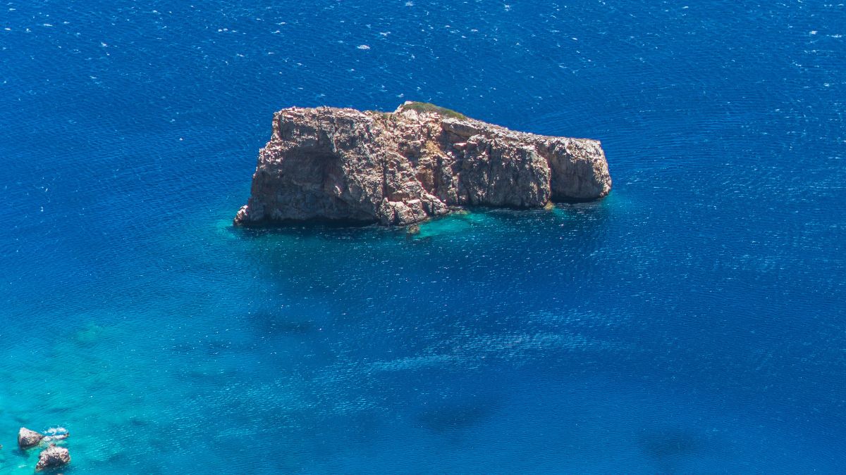 A rocky island emerges from vibrant blue waters, surrounded by lighter shades of turquoise. The calm sea reflects sunlight, creating a shimmering effect.