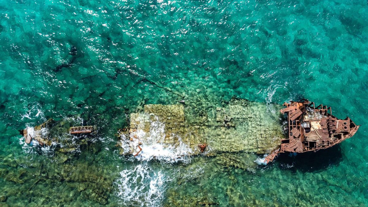 Aerial view of a shipwreck partially submerged in crystal-clear turquoise waters, with surrounding rocky seabed and gentle waves lapping at the remains.