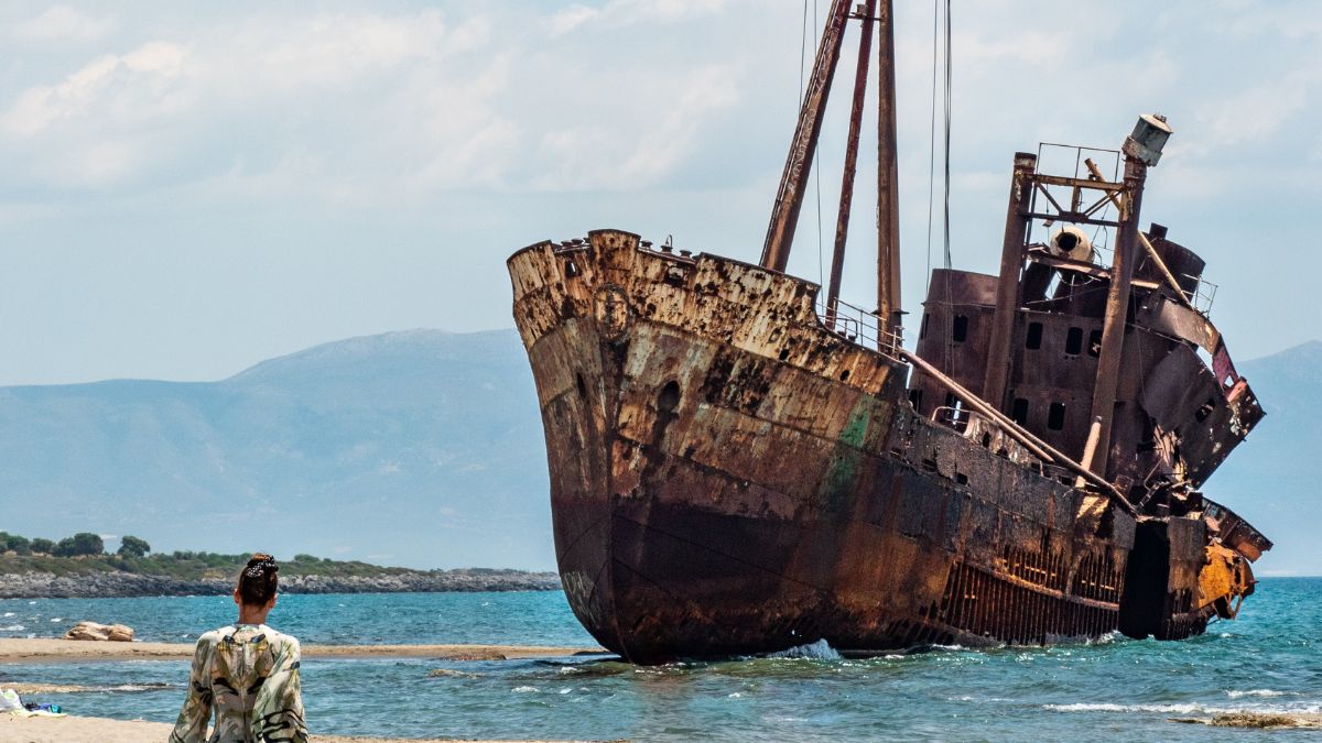 A rusted shipwreck stands beached on a shore, with a person standing in the foreground gazing at it. The background features a calm sea and distant mountains under a partly cloudy sky.
