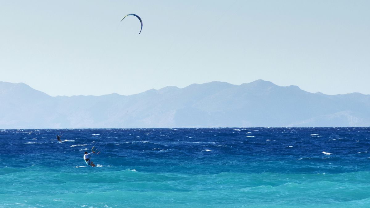A kite surfer glides across vibrant blue waters, with mountains in the background against a clear sky.