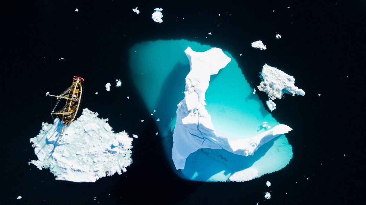 A wooden ship anchored near a large, white iceberg surrounded by turquoise water. Smaller ice fragments float nearby in the deep blue sea.