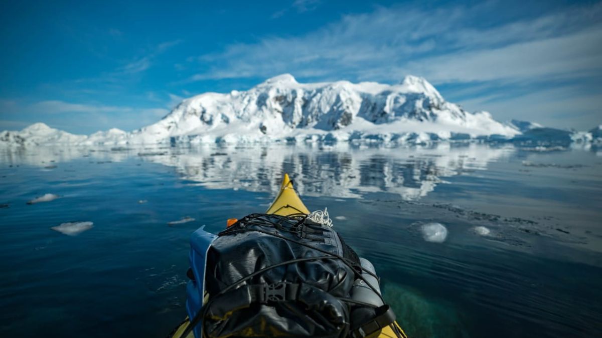 A yellow kayak floats on calm, icy waters, with snow-capped mountains in the distance under a clear blue sky. The reflection of the mountains and ice is visible in the water.