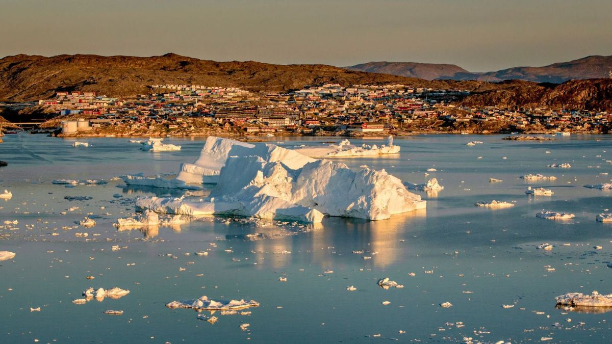 A serene landscape featuring large icebergs floating in calm waters, with a colorful town nestled against rocky hills in the background. The sunlight illuminates the ice, creating a tranquil scene of nature and human settlement.
