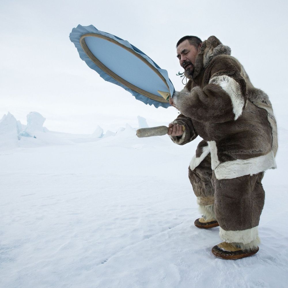 A man dressed in traditional fur clothing plays a drum on a snowy, icy landscape, with frozen formations in the background.