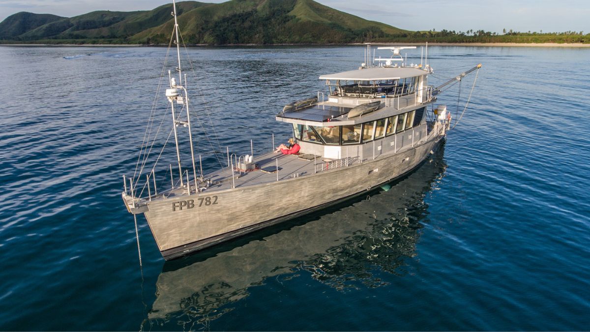 A sleek gray motorboat sails on calm waters with a mountainous shoreline in the background. The sun reflects off the surface, illuminating the boat's features.