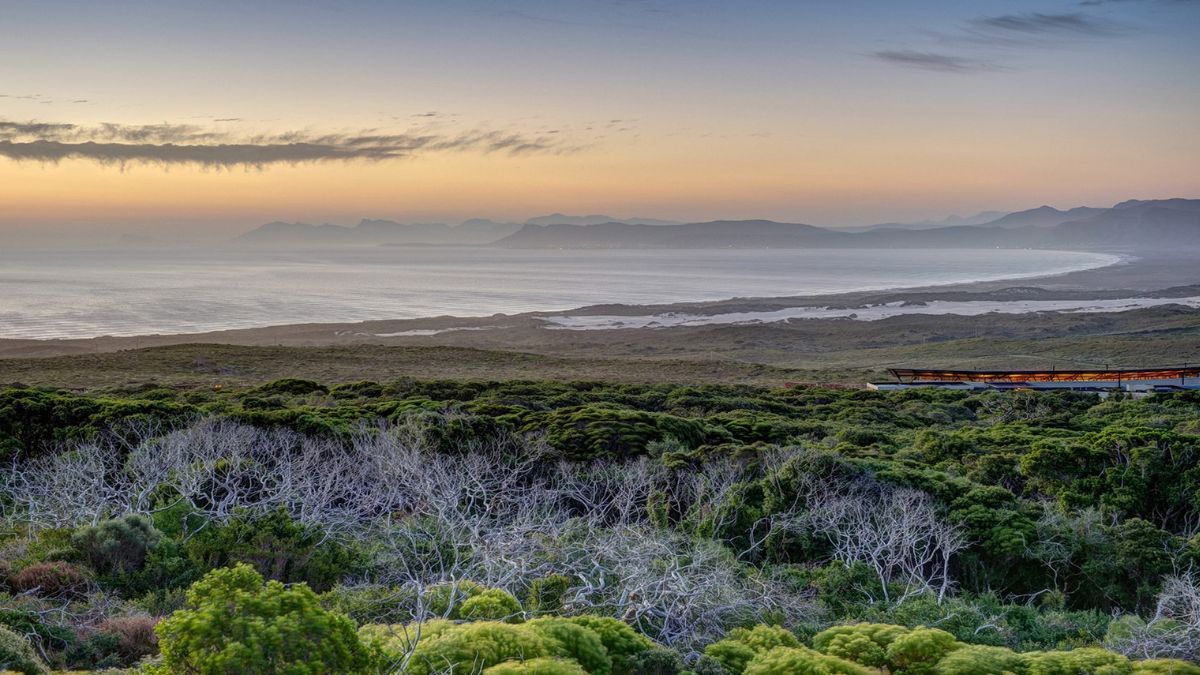 A serene coastal landscape at sunset, featuring rolling hills, a sandy beach, and gently lapping waves. The sky is painted with soft pastel colors, transitioning from blue to orange as the sun sets behind distant mountains. Rich greenery lines the foreground, adding depth to the scene.