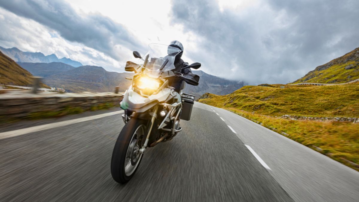 Person riding a silver touring motorcycle with headlights on along a paved mountain road under a cloudy sky.