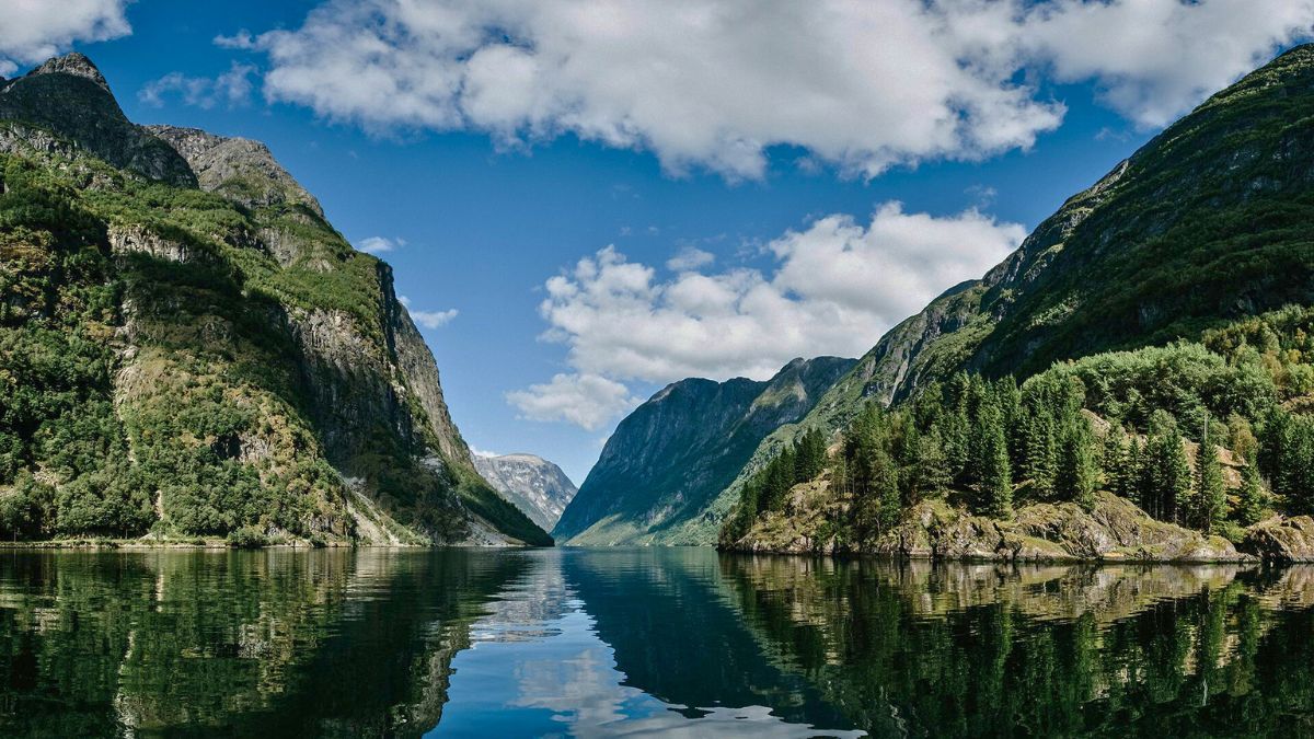 A serene fjord surrounded by towering green mountains under a partly cloudy blue sky. The calm water reflects the landscape, creating a mirror-like effect.