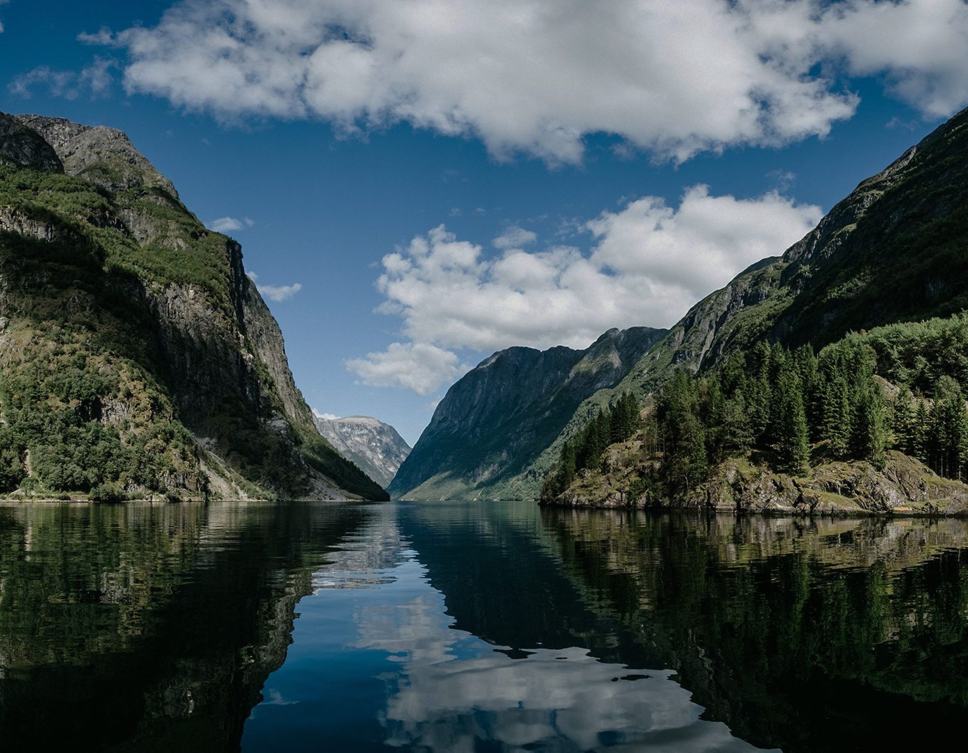 A serene fjord surrounded by towering mountains and lush greenery, with calm water reflecting the dramatic landscape under a partly cloudy sky.