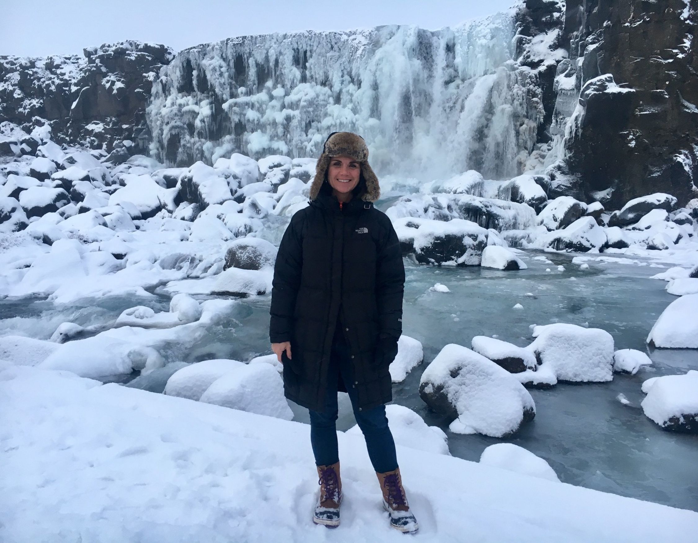 A Pelorus Travel expert, Gwen, in a black parka and fur hat stands in a snowy landscape in front of a partially frozen waterfall.