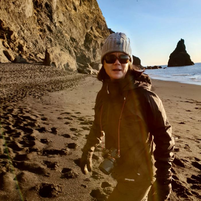 A woman wearing sunglasses, a knitted beanie, and a warm jacket stands on a sandy beach. She has a camera around her neck and is smiling at the camera, with rugged cliffs and ocean waves in the background.