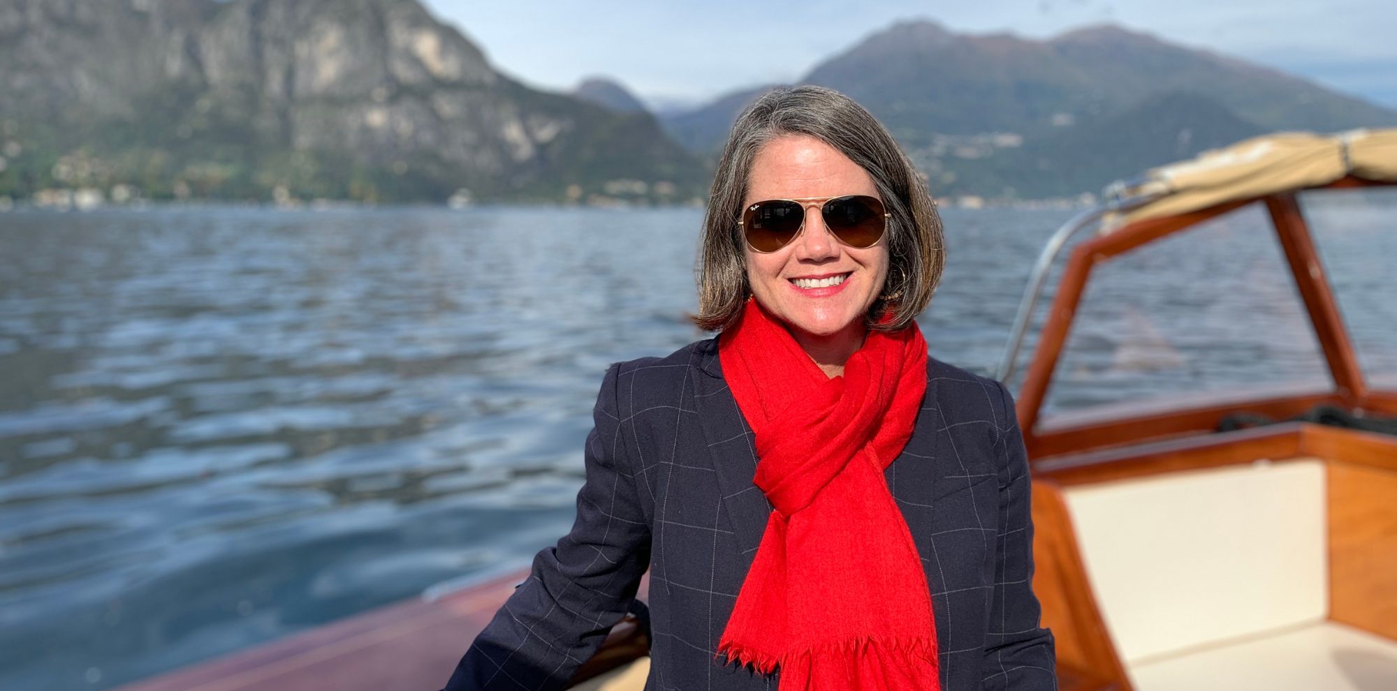 A woman with sunglasses and a red scarf stands on a boat, smiling, with mountains and calm water in the background.