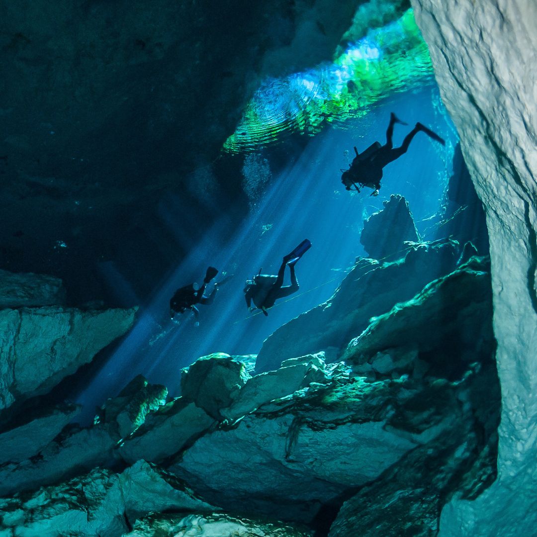 Three scuba divers explore an underwater cave, surrounded by crystal-clear blue water and dramatic rock formations. Rays of sunlight penetrate the surface, creating a serene and mystical atmosphere.