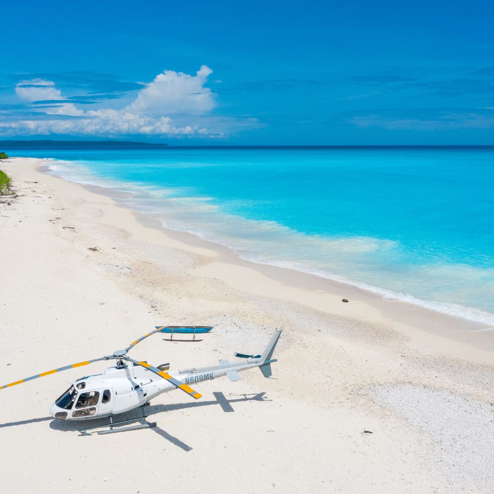 White helicopter parked on a pristine white sand beach next to turquoise ocean water.