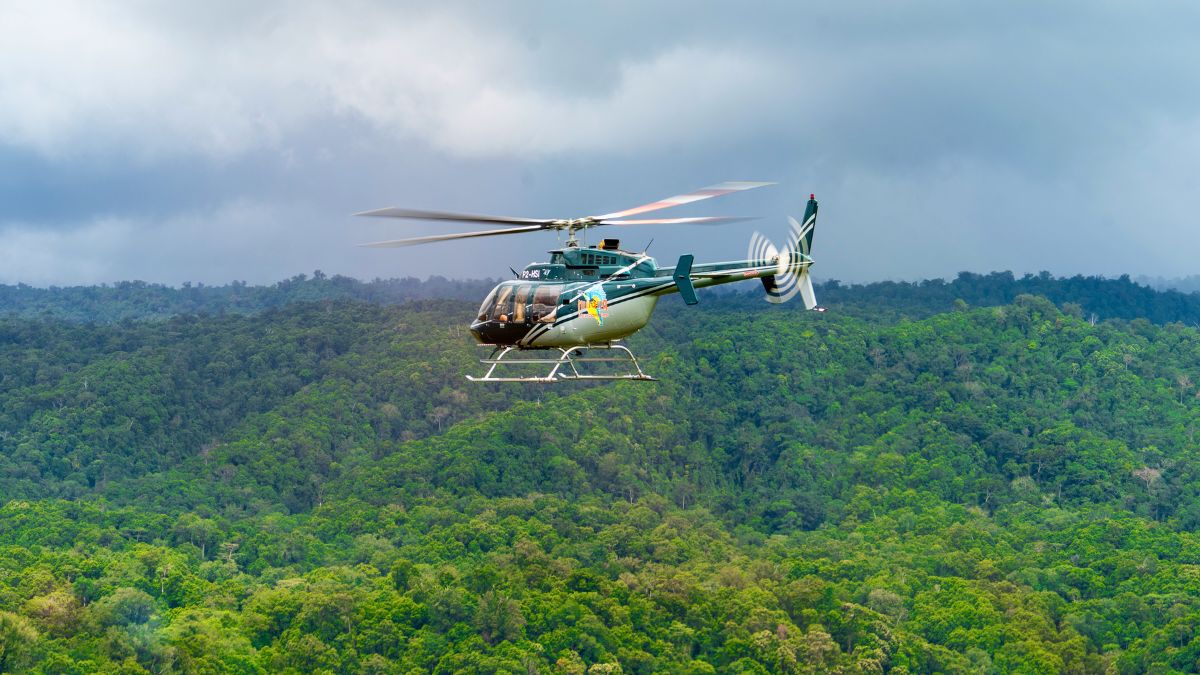 A green and white helicopter flies over a vast, dense tropical rainforest under a grey, overcast sky.