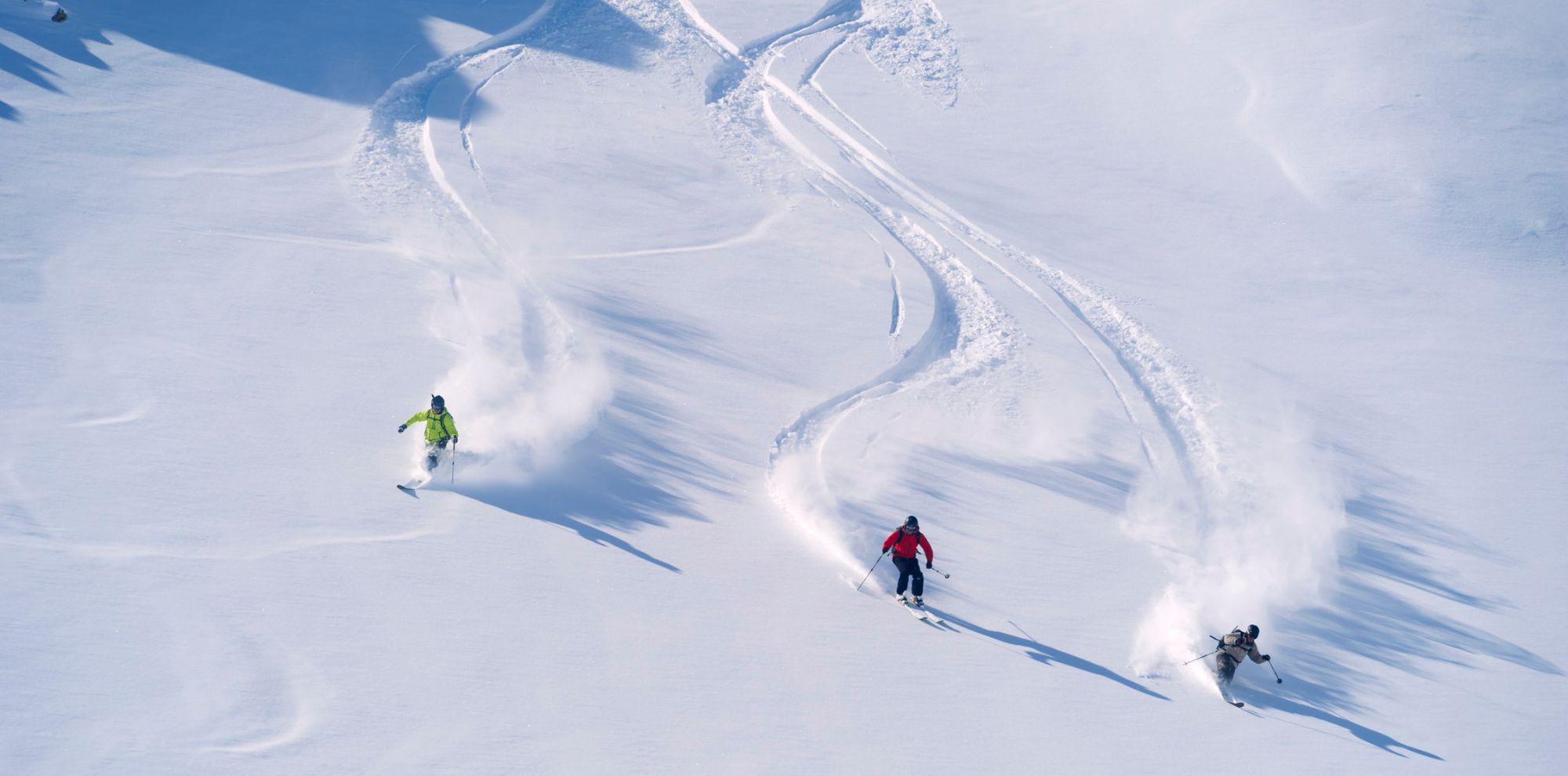 Aerial view of three skiers carving through deep white powder, leaving curved tracks on a bright snow-covered slope.
