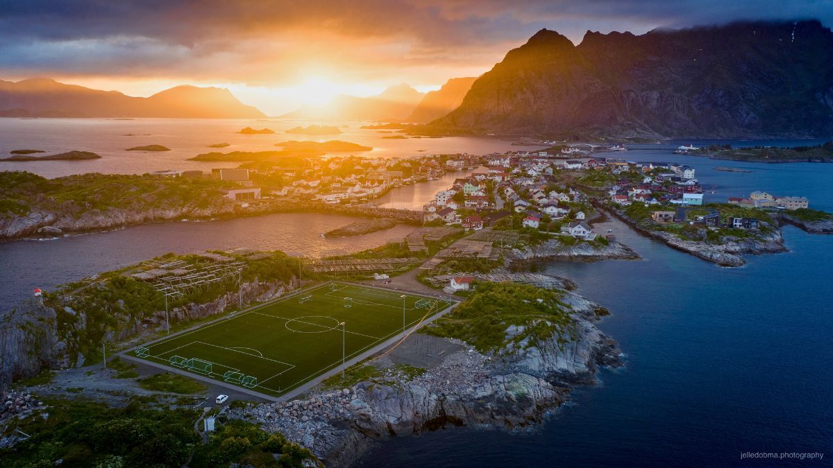 Aerial view of a soccer field on a rocky island village in Norway during a golden sunset over the mountains and ocean.