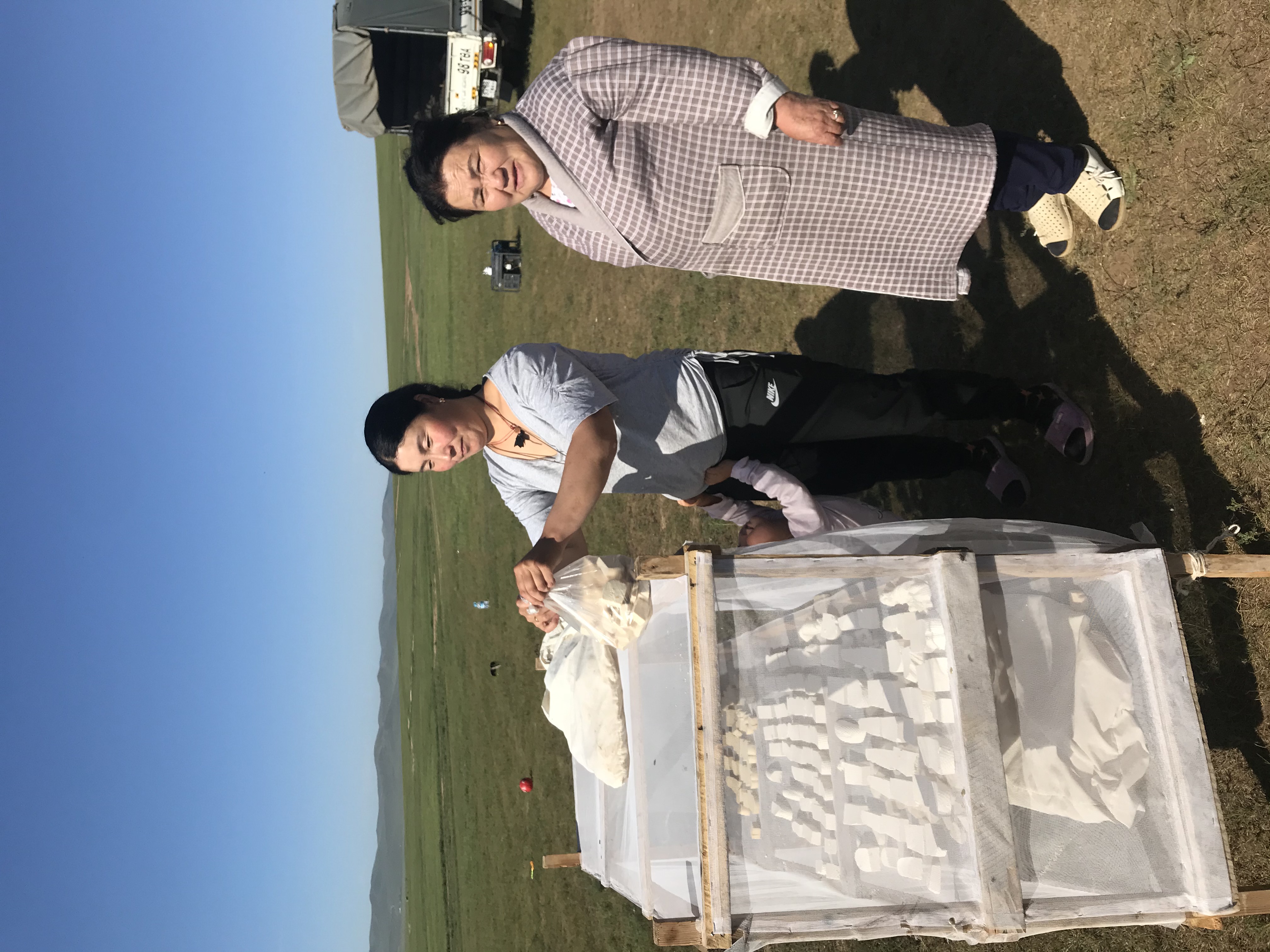 Two women are outdoors in a grassy landscape under a clear blue sky. One woman, dressed in athletic wear, is placing items into a wooden display covered with mesh. The other woman, wearing a patterned robe, observes with a smile. A child can be seen partially behind the display.
