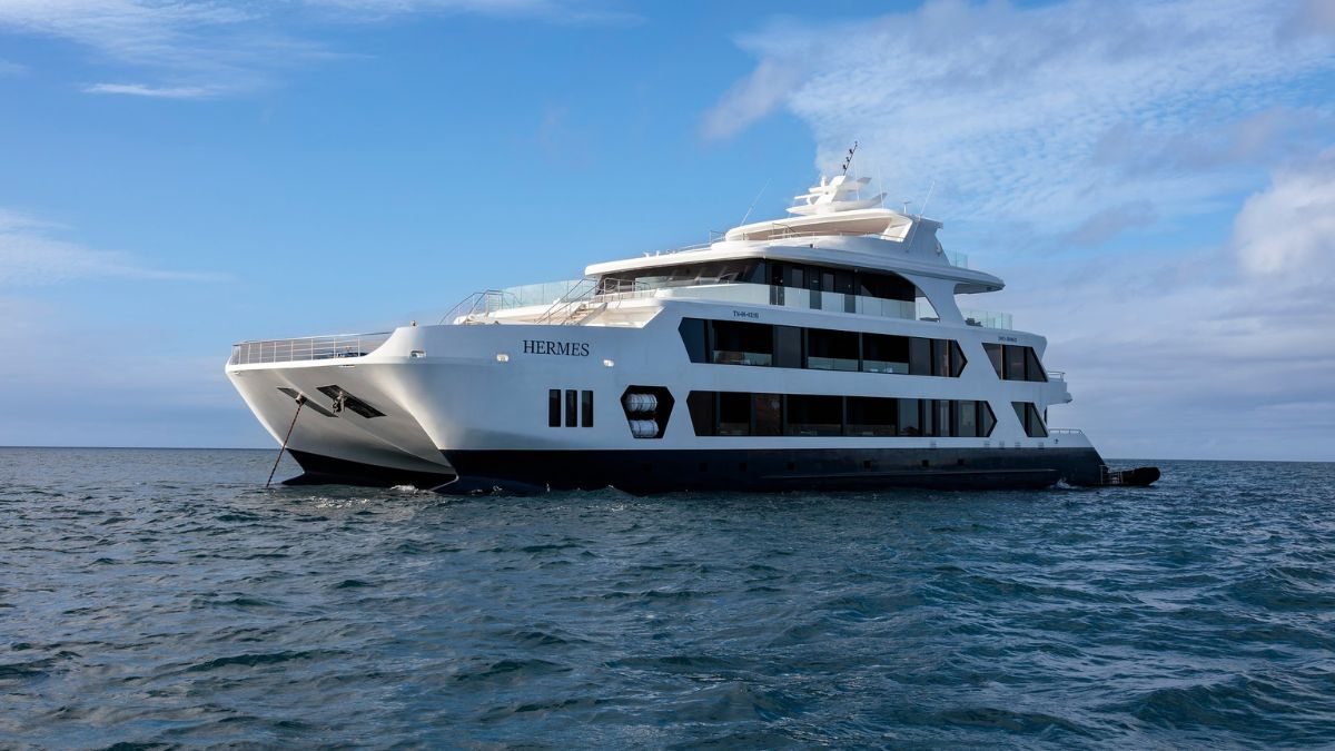 A white luxury catamaran yacht named HERMES with dark windows anchored on the ocean under a cloudy sky.