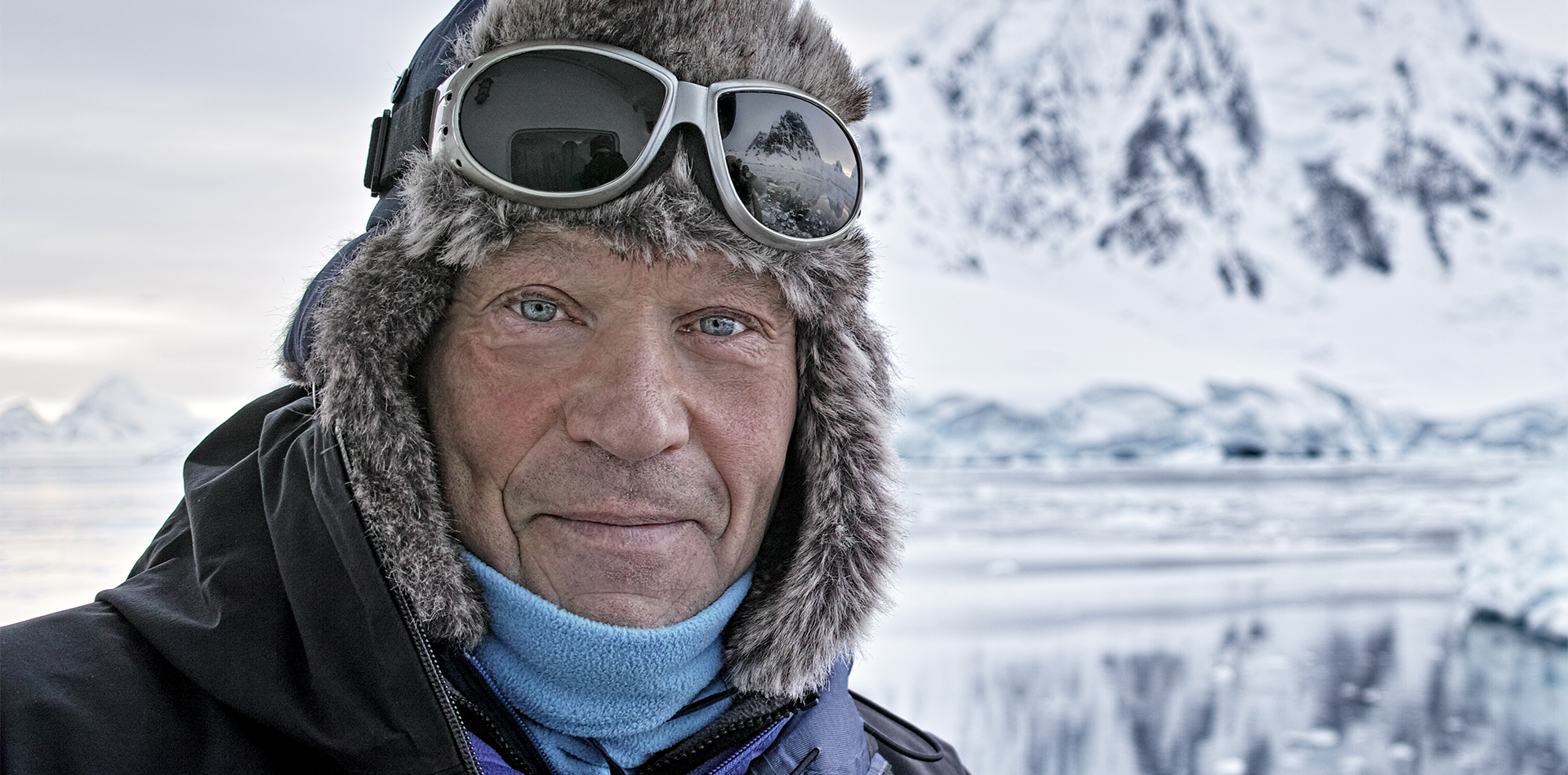 An older man with blue eyes and a serious expression wears a fur-lined hat and goggles. He stands against a snowy landscape with ice formations and mountains in the background.