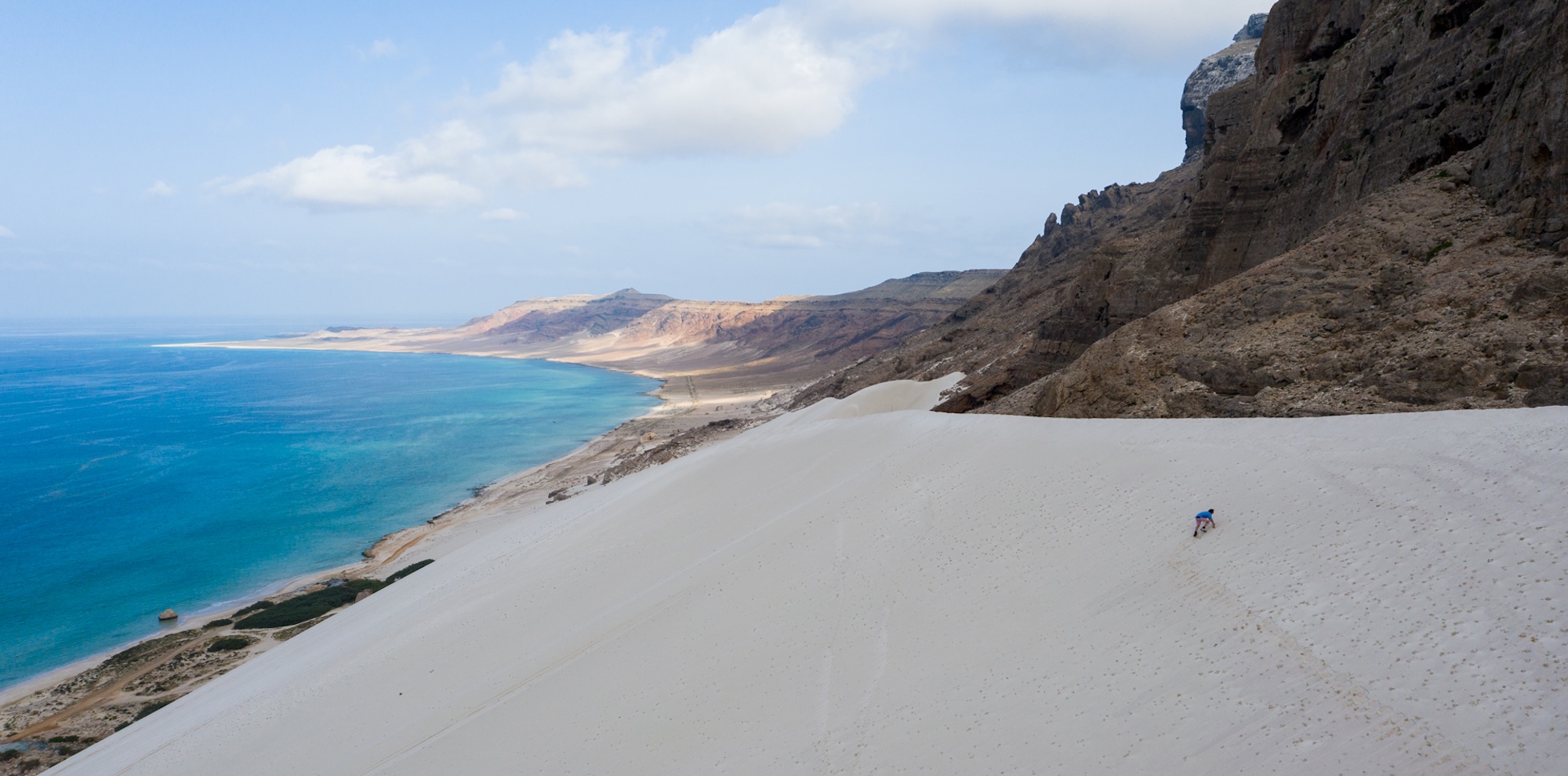 A vast sandy dune slopes down towards a turquoise sea, with rugged cliffs and colorful hills in the background. The sky is partly cloudy, adding depth to the serene landscape. A small figure is seen climbing the dune, highlighting the scale of the scene.