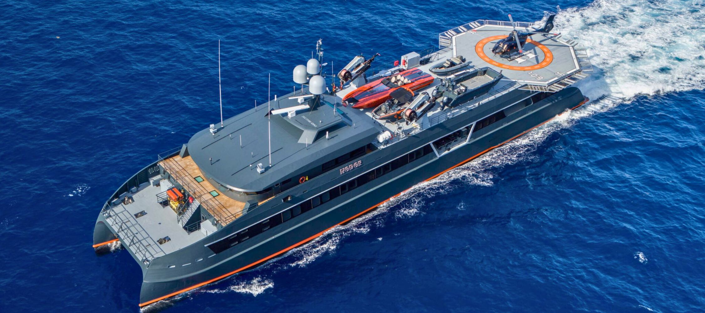 A large motor yacht cruises through deep blue ocean waters, displaying a sleek design with a spacious deck. Various small boats and equipment are visible on the deck.