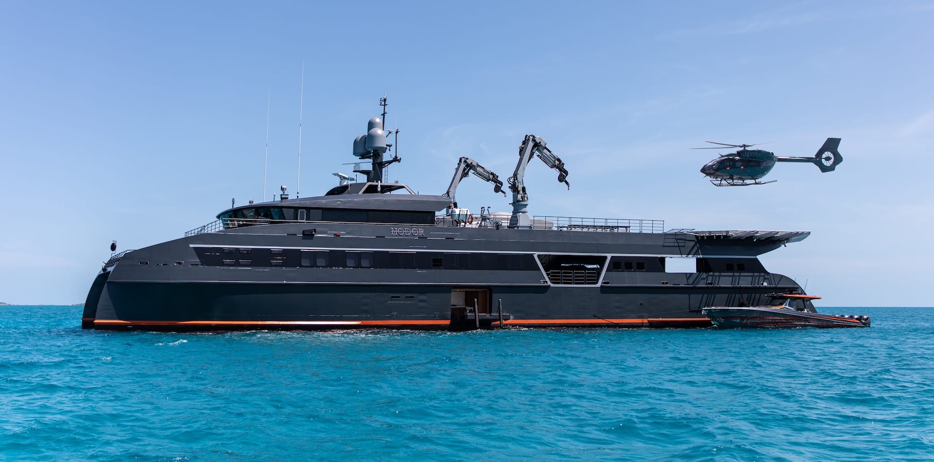 A large, sleek black yacht is anchored in clear blue waters, featuring multiple robotic arms and a helicopter hovering above it. The yacht has a modern design and an extended platform at the rear.