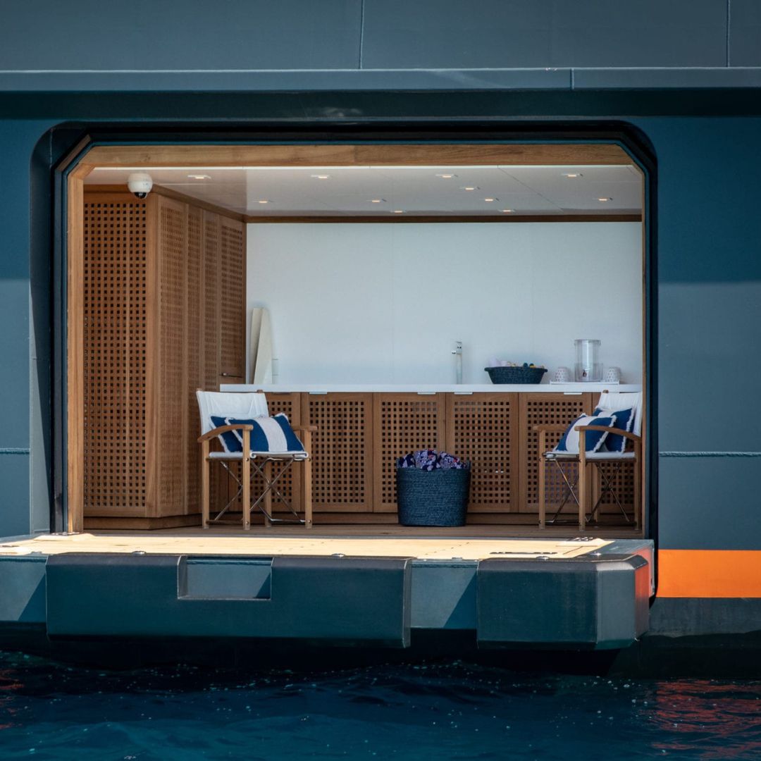 A modern boat interior featuring light wood cabinetry and a minimalistic design. Two white and blue chairs sit in front of a counter with a basket of flowers. The scene opens to the water, highlighting a serene nautical atmosphere.