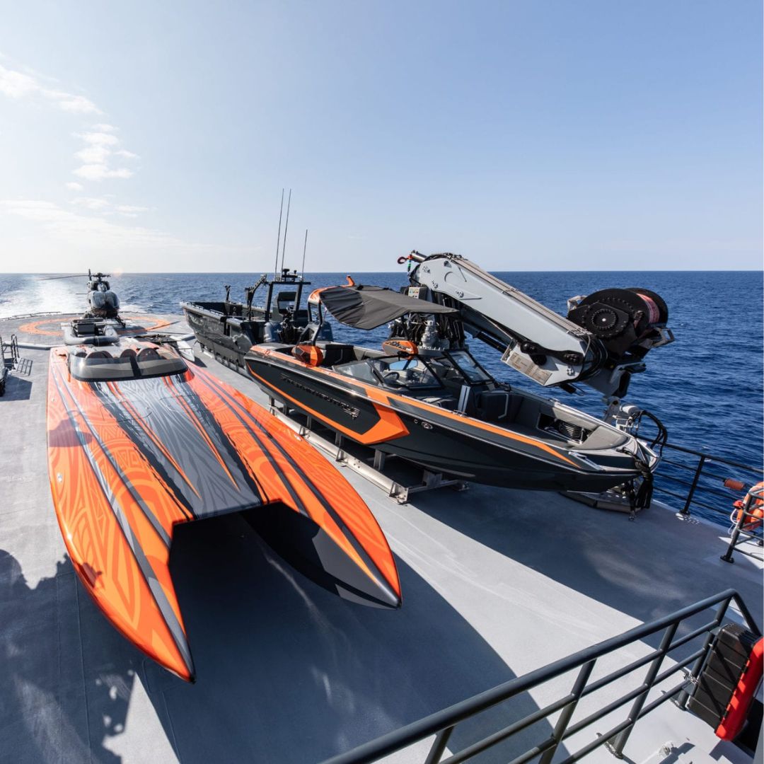 Two high-performance boats in bright orange and gray colors are secured on the deck of a ship, with the ocean visible in the background. One is partially elevated on a lift, and sunny skies stretch above.