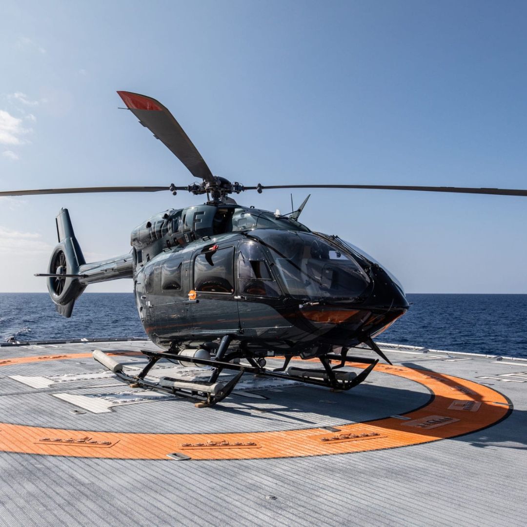 A military helicopter is stationed on a helipad over the ocean, with clear blue skies in the background. The helicopter is sleek and black, showcasing its rotors and landing gear.