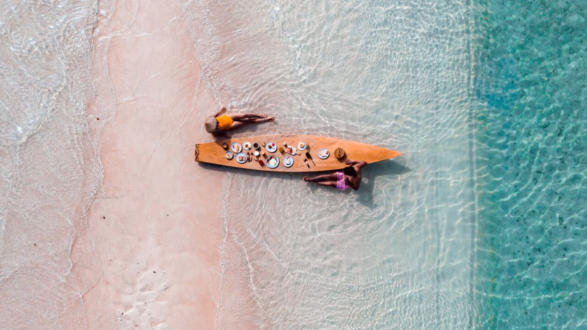 Overhead view of two people relaxing in shallow turquoise water on a pink sand beach next to a surfboard holding a breakfast spread.