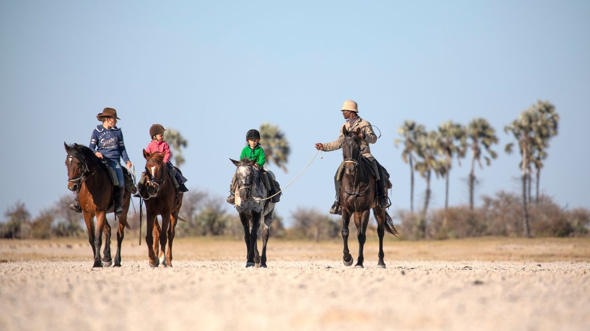 A group of four people, including two children, riding horses across a dry, flat landscape under a clear blue sky.