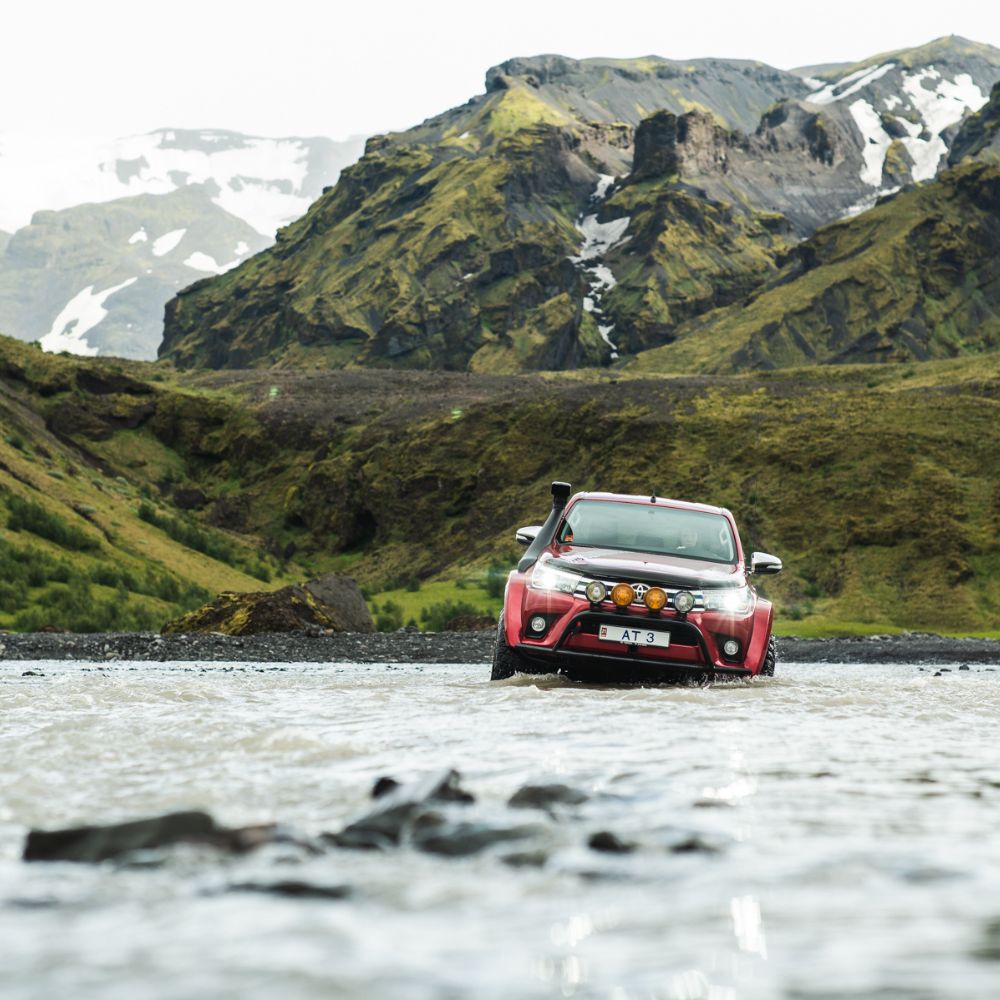 Red off-road vehicle driving through a shallow river in a rugged mountain landscape.