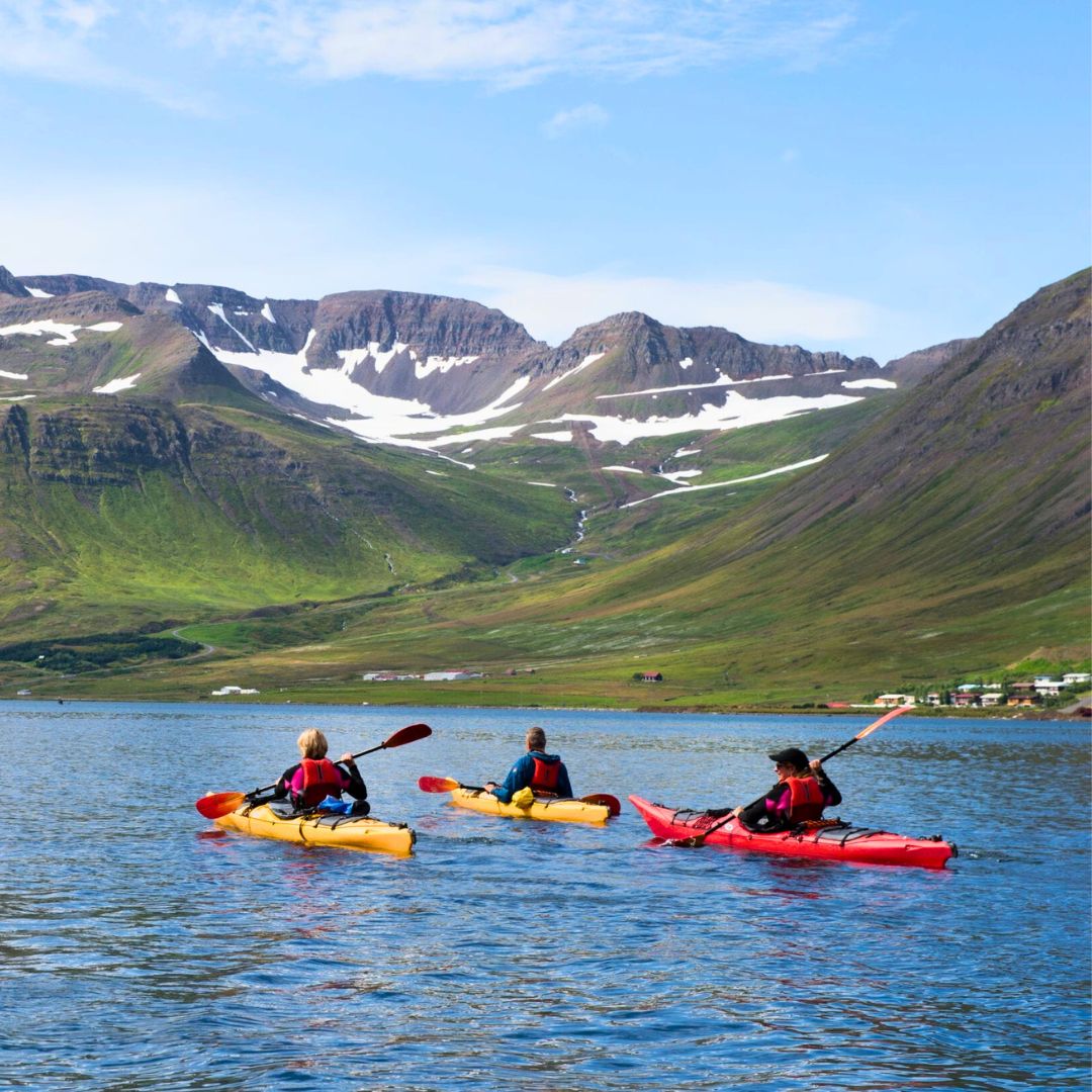 Three people in yellow and red kayaks paddling on a blue bay with green, snow-streaked mountains in the background.