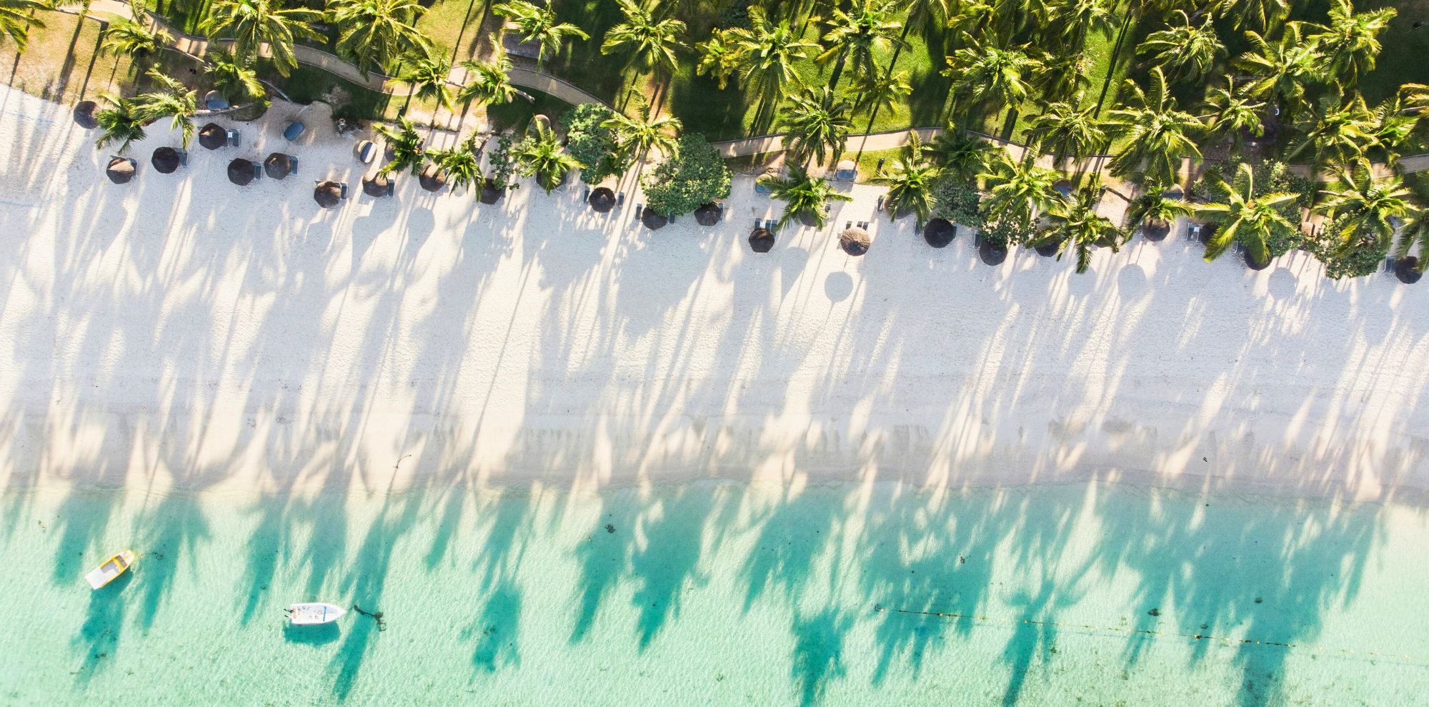 Aerial view of a white sand beach lined with palm trees casting long shadows onto turquoise water with two small boats.